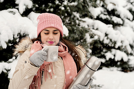 喝温水穿着温暖冬衣的漂亮女人 拿着热温水和在雪天户外喝热茶或咖啡 女性 树背景
