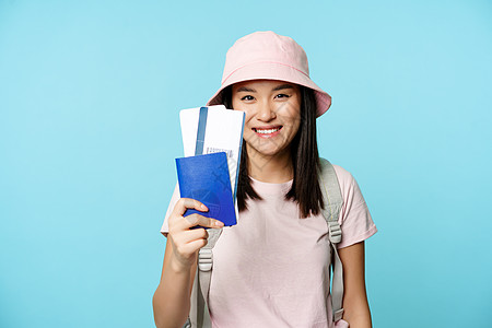 旅游和游客的概念 微笑快乐的亚洲女孩旅行者 戴着巴拿马帽 出示带机票的护照 预订旅行 蓝色背景图片