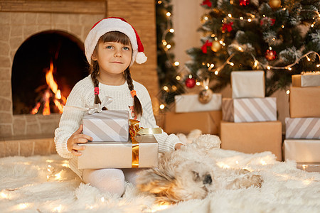 沉思的小孩坐在地板上 看着别处 拿着带丝带的礼盒 想象她的礼物 在装饰着花环 圣诞树和壁炉的客厅里摆姿势图片