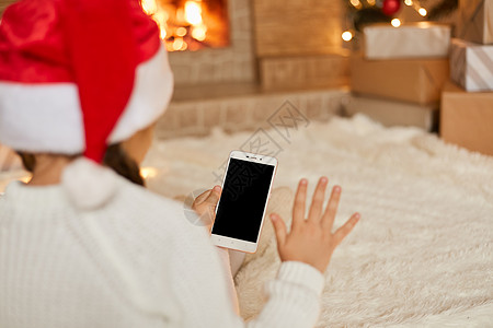 小女孩看着空白的手机屏幕 挥舞着手 背影是戴着圣诞帽和白色套头衫的女孩坐在壁炉旁地板上的柔软地毯上图片