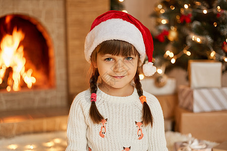快乐微笑的小女孩在装饰的客厅里摆姿势 孩子直视相机 扎着辫子 穿着白色套头衫和红色圣诞老人帽子 在圣诞树和壁炉旁摆姿势图片
