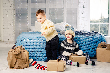 在阳光明媚的日子里 孩子们的兄弟姐妹坐在床边卧室的地板上 在圣诞装饰的背景下拿着礼物 穿着温暖的针织羊毛衣帽和彩色连指手套 快乐图片