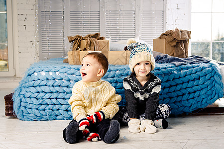 在阳光明媚的日子里 孩子们的兄弟姐妹坐在床边卧室的地板上 在圣诞装饰的背景下拿着礼物 穿着温暖的针织羊毛衣帽和彩色连指手套 装饰图片