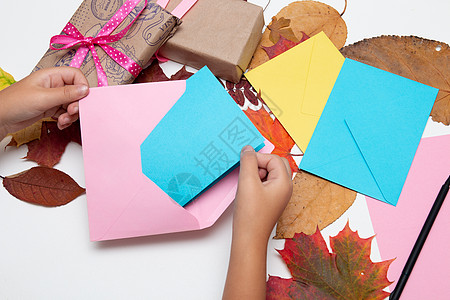 孩子们的手把卡片放在信封里 鲜艳的颜色 带漂亮丝带的礼物 节日问候 感恩节 生日 白桌上的秋叶 复制空间顶视图 粉色 黄色 蓝色图片