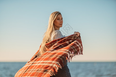 迷人的金发白种女人在日落时分在海滩上享受时光 裹着毯子走着 看着旁边 背景是落日的天空和大海 海滩度假 呼吸 水图片