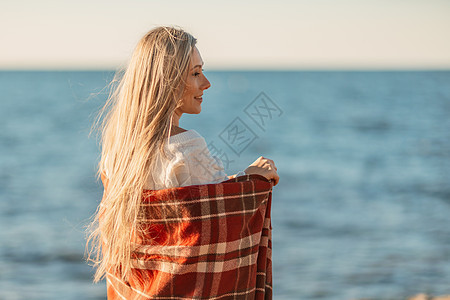 迷人的金发白种女人在日落时分在海滩上享受时光 裹着毯子走着 看着旁边 背景是落日的天空和大海 海滩度假 户外 晴天图片