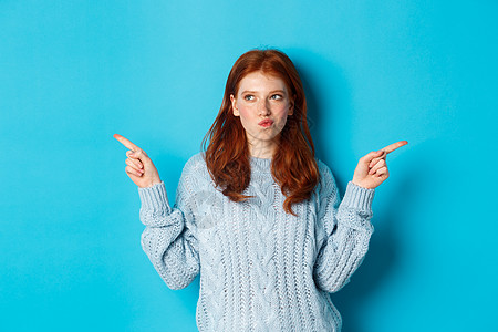 针织毛衣寒假和人们的观念 深思熟虑的红发女孩做出决定 手指横着 在两种方式之间做出选择 站在蓝色背景上 舒适 红色的背景