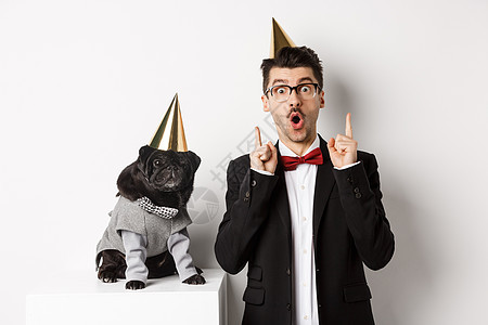 穿着派对锥狗和站在快乐的主人身边 男人在复制空间指着手指 庆祝生日 白背景等可爱的黑小狗 衣服 锥体图片