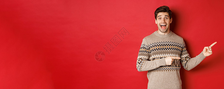 圣诞节庆祝 冬季假日和生活方式的概念 穿着Xma毛衣的帅哥指对手指 笑笑惊讶 在红色背景下展示新年宣战活动 笑声 冬天 快乐的背景图片