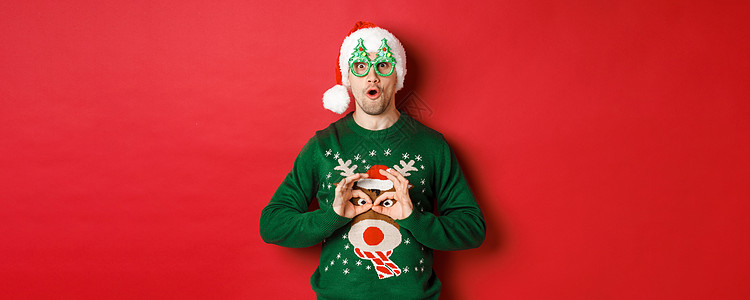穿着圣塔帽和派对眼镜的无忧无虑英俊男子肖像 取笑他的圣诞节毛衣 在红色背景下看着快乐 男生 成人背景图片