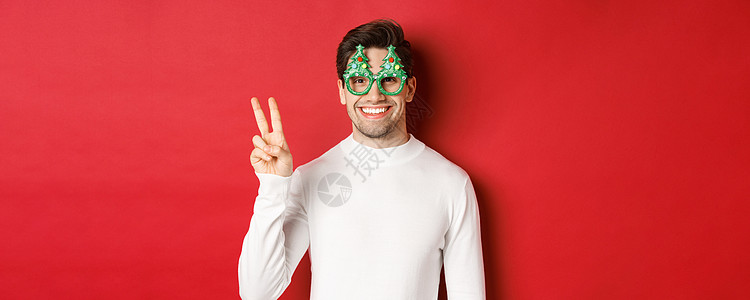 穿着白色毛衣和政党眼镜的英俊男子照片 展示和平标志和微笑 祝愿圣诞快乐 站在红色背景上 工作室 庆祝背景
