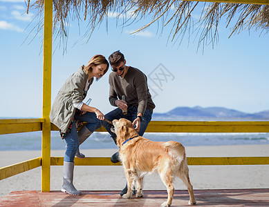 狗的高清素材年轻夫妇和一条狗在沙滩上 晴天 假期 海洋 空的背景