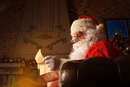 快乐圣诞老人的画像坐在他在家里的房间里靠近圣诞树 阅读圣诞信或愿望清单 希望 男性背景图片