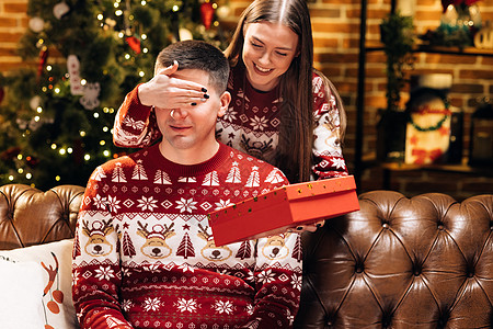 微笑的 30 多岁年轻女性遮住微笑的好奇丈夫的眼睛 在装饰过的节日树附近送上装有圣诞礼物的包装盒 新年寒假家庭庆祝活动 假日奇迹图片