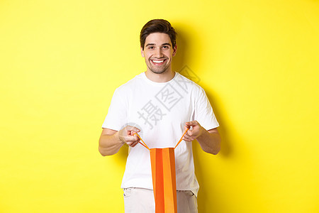 快乐的青年男子打开购物袋 现时打开 微笑着镜头 站在黄色背景上 时尚 广告技术高清图片素材