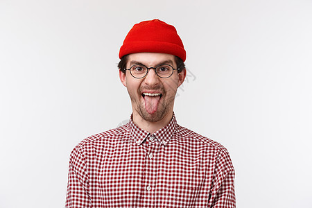 特写肖像滑稽可爱的留着胡子的嬉皮士 戴着红色的帽子和眼镜 笑着开玩笑地吐舌头 玩得开心 表现得像个孩子气的站在白色背景上的快乐图片