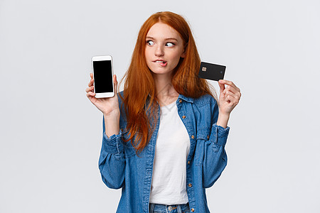 女孩考虑以超低折扣购买东西 看着智能手机 购物移动应用程序 持有信用卡 咬唇急切购买的红发女性购物狂图片