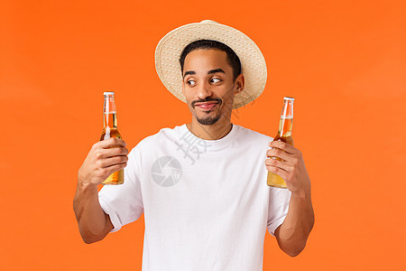 戴着帽子的快乐 微笑和开朗的白种人在热带国家旅行时放松 用冷可口的饮料冷却自己 拿着两瓶啤酒 站立橙色背景图片