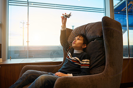 在窗户前的男孩帅气的白人青春期男孩坐在椅子上玩玩具飞机 全景窗户旁俯瞰日落时分的跑道 同时在机场出发航站楼等待登机背景
