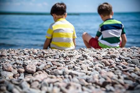 两个白人男婴坐在海滩上 孩子们穿着裤子 背靠在海边的岩石鹅卵石上 和孩子一起出海度假 男生 太阳图片