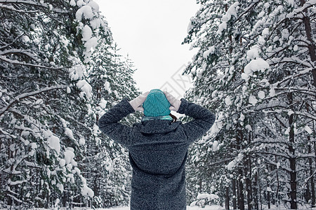 身着灰色大衣和浅蓝色编织帽子的女孩 双手站在美丽的冬季雪林中的头顶上图片