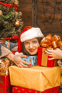 戴着红色圣诞帽的可爱的年轻女人拿着一打圣诞多色礼物 装饰圣诞树 墙上的电动花环 灯 特写 木背景图片