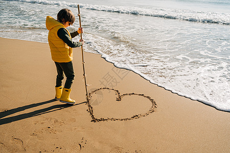 穿着黄色橡胶靴的男孩在沙滩上的沙子上画心形 学校的孩子在秋冬海上接触水 孩子在岸边玩海浪 春假假期概念 棕榈 海滨图片