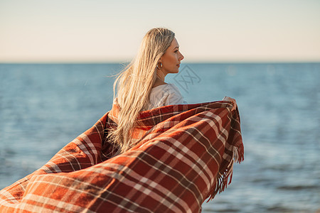 迷人的金发白种女人在日落时分在海滩上享受时光 裹着毯子走着 看着旁边 背景是落日的天空和大海 海滩度假 真诚 海岸线图片