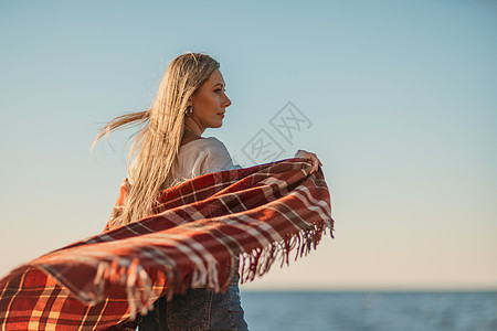 迷人的金发白种女人在日落时分在海滩上享受时光 裹着毯子走着 看着旁边 背景是落日的天空和大海 海滩度假 女孩 晴天图片