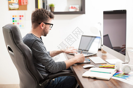 一个英俊的年轻男子带着眼镜在办公室工作同时使用两台电脑的侧面图片