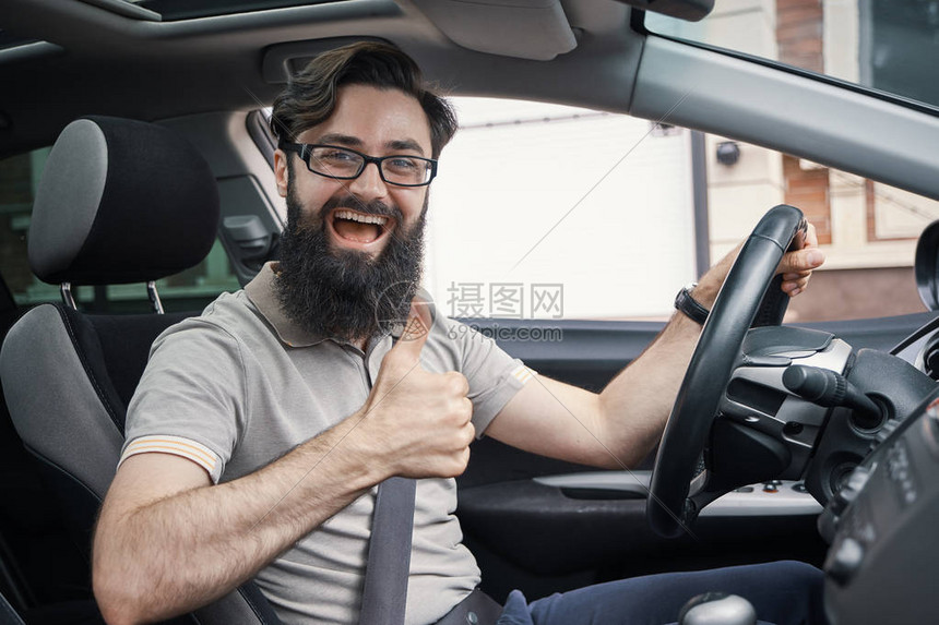 男司机开心地笑着竖起大拇指驾驶着城市建筑在背景中的跑车英俊的年轻人对他的新车感到兴奋图片