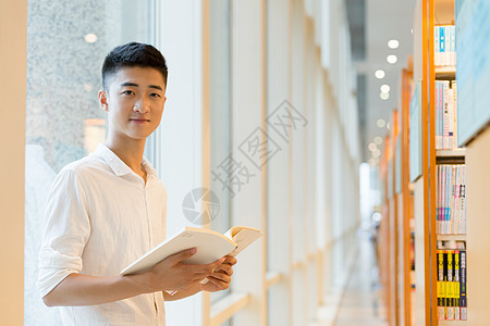 拿着书的学生站在图书馆书架旁看书的帅气男高中生同学背景