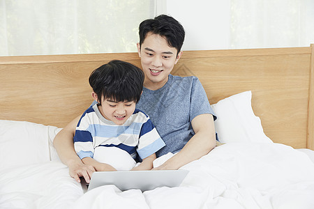 年轻爸爸陪儿子在床上使用笔记本电脑图片