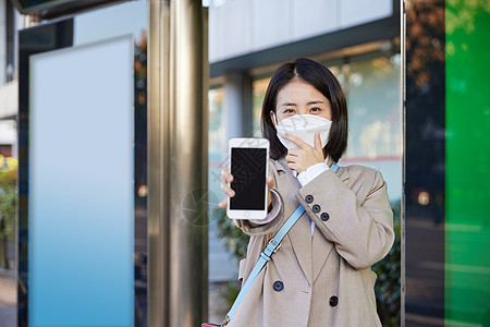 戴口罩在公交站台等待公交车的女性展示手机背景图片