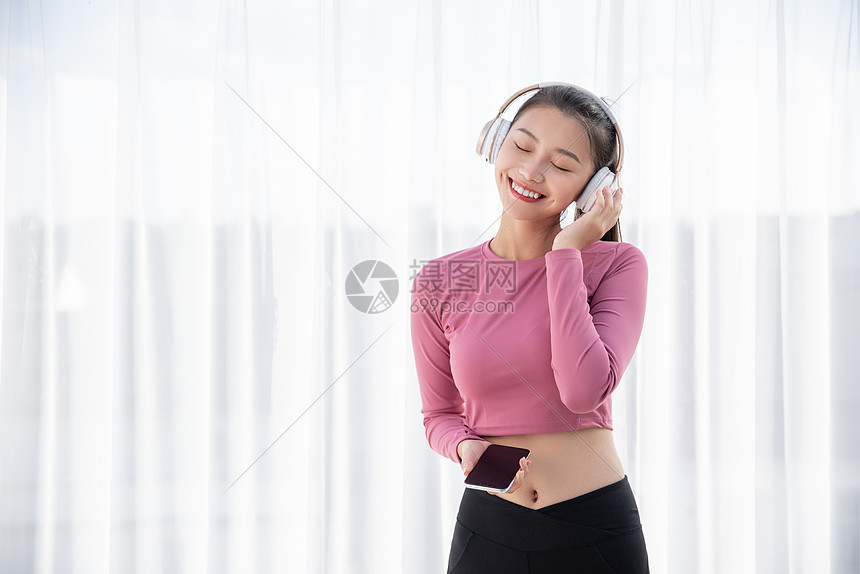 穿着运动装的年轻美女在窗边戴耳机听音乐图片