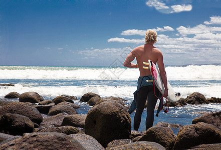 年轻的男性冲浪者站在海滩岩石上向远外看图片