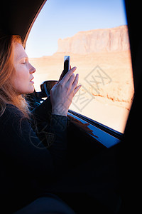 在美国犹他州古迹谷汽车窗口用智能手机上拍摄照片的女性图片