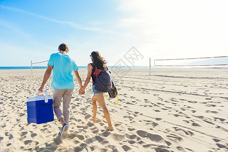 年轻夫妇在海滩上行走拿着凉爽的盒子后视图片