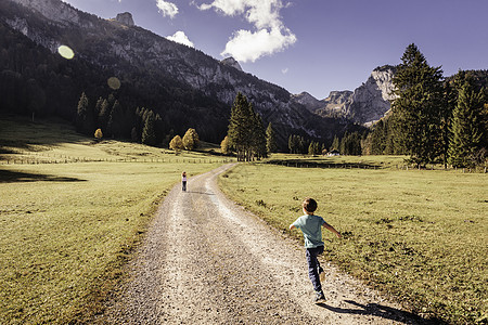 在德国巴伐利亚的山谷风景中男孩在泥土路上向女孩跑去的后视角图片