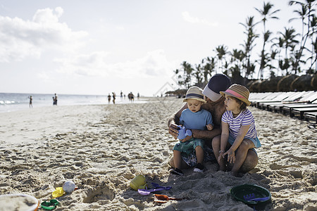 父亲和儿子父亲坐在沙滩上与小儿子和女坐在一起背景