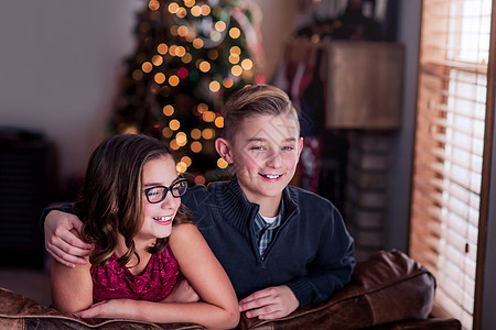 在圣诞树背景下靠在沙发上笑着的兄弟姐妹们肩并肩高清图片素材