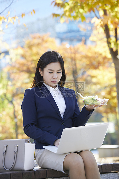 长凳上拿着沙拉使用笔记本电脑的女性图片