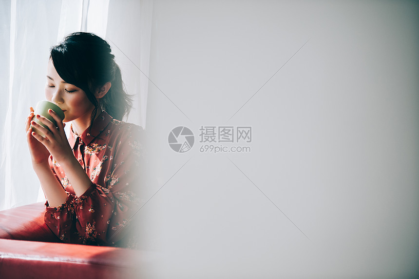 穿着红裙的女孩坐在靠窗的沙发上喝咖啡图片
