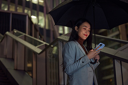 边走路边玩手机城市夜晚撑着伞的商务女性背景