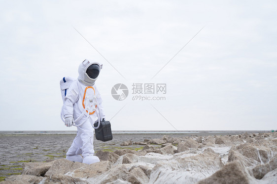 穿着宇航服的男性拿手提箱行走在海边图片