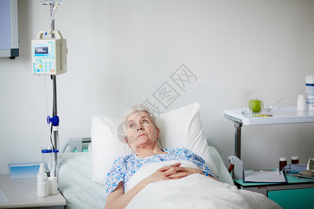 生病的老年妇女在得到瓶子滴水处理后躺在医院床上休息图片