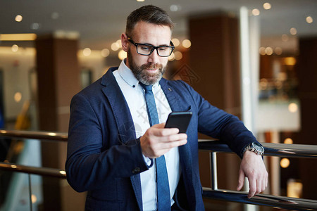 身着优雅正装和眼镜靠在办公楼栏杆上阅读智能手机短信的成功图片