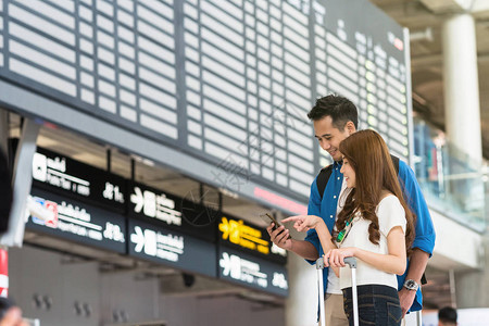 亚洲夫妇旅客使用智能手机在现代机场的航班信息屏幕上办理登机手续图片