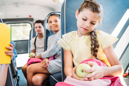 带着背包和苹果坐校车的漂亮小女学生与背景模糊的同学一图片