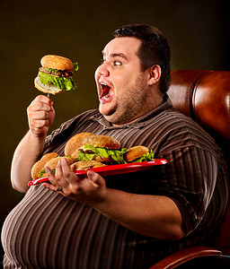 吃快餐汉堡包的胖子的饮食失败快乐的微笑超重的人通过在叉子上吃巨大的汉堡包而破坏了健康食品他在皮椅上提着背景图片